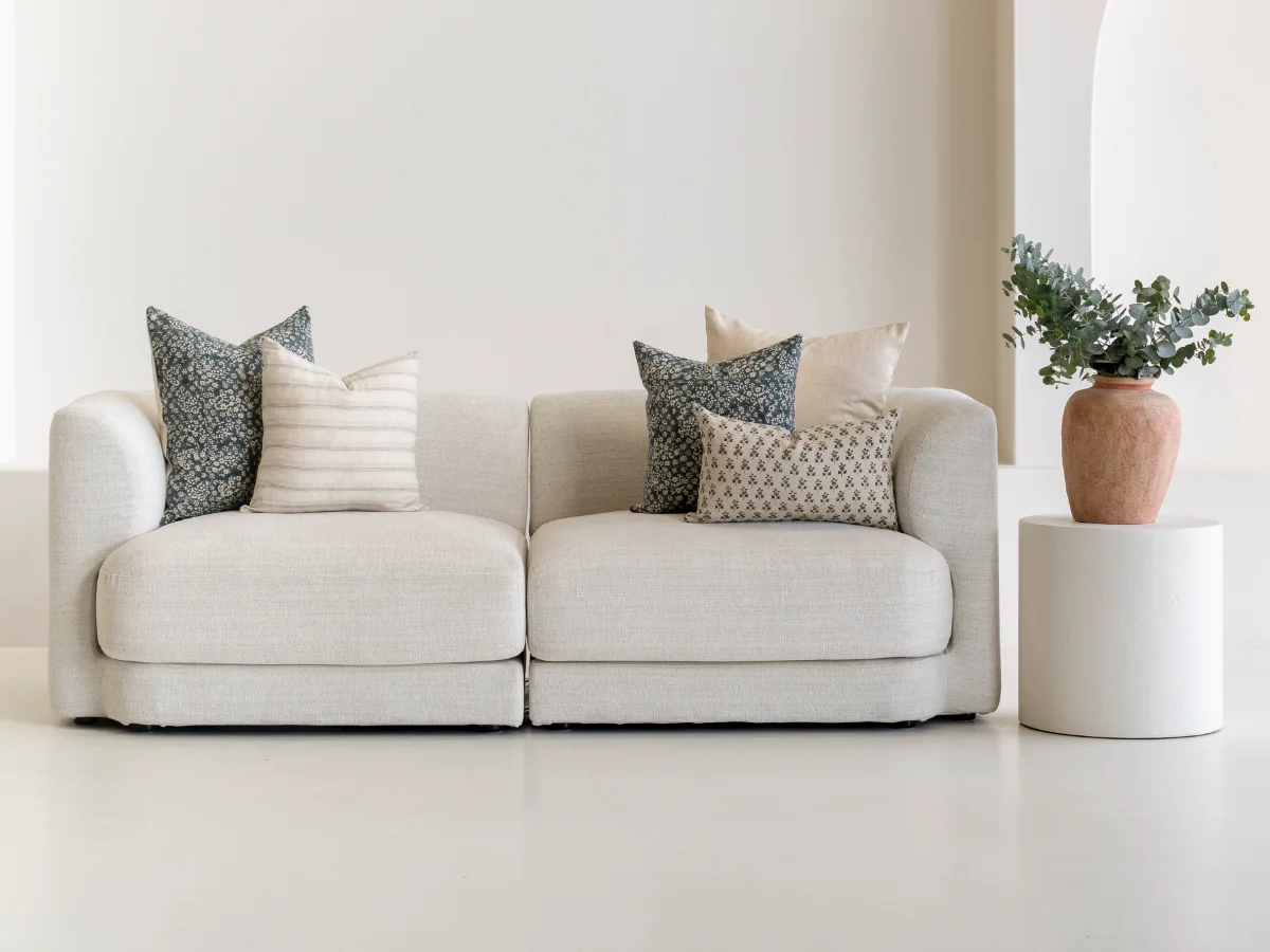 Five patterned arranged cushions on a sofa with a plant resting on a side table.