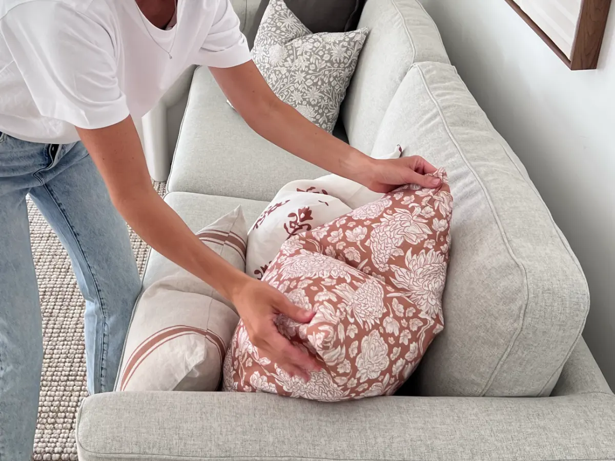 A lady is puffing a cushion to arrange it as part of a set on a grey sofa.