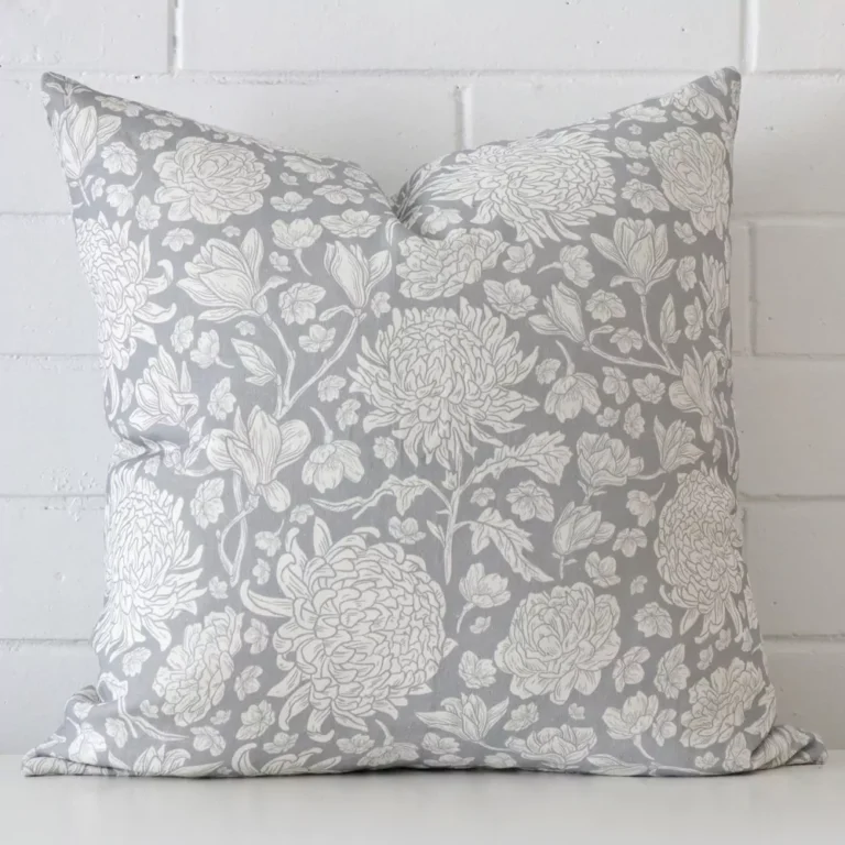 An attractive floral linen cushion in front of a white brick wall. It has a square shape and is ligt blue in colour.