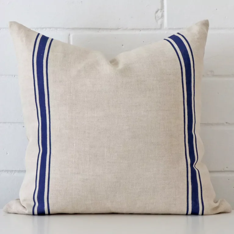 Bold square blue cushion positioned in front of white brickwork. Its striped style pops on the linen fabric.