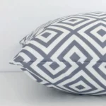 Grey geometric cushion cover laying sideways against white wall. The square size and outdoor material are shown highlighting the seams.