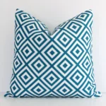 Teal geometric cushion cover in front of a white wall. It has a square size and is made from an outdoor material.