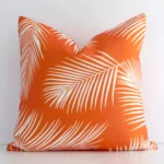 White wall with a palm-designed orange cushion laying against it. It has a distinctive outdoor fabric and has a square shape.