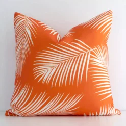 White wall with a palm-designed orange cushion laying against it. It has a distinctive outdoor fabric and has a square shape.
