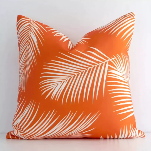 White wall with a palm-designed orange cushion laying against it. It has a distinctive outdoor fabric and has a square shape.