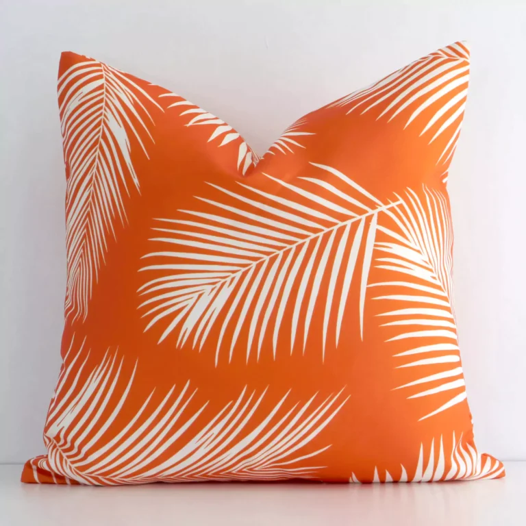 White wall with a palm-designed orange cushion laying against it. It has a distinctive outdoor fabric and has a square shape.