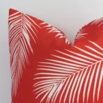 Enlarged shot of the corner of this square palm designed cushion cover in red colour is shown against a brick wall. The image shows the quality and craftsmanship of the outdoor material.