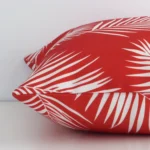 A red palm styled cushion arranged sideways in front of a wall. The square shape and outdoor fabric are shown and the seams are clearly visible.