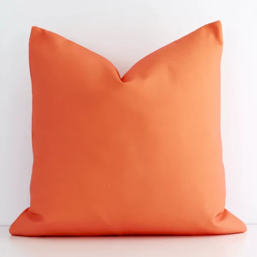 An attractive solid coloured outdoor cushion in front of a white brick wall. It has a square shape and is coral in colour.