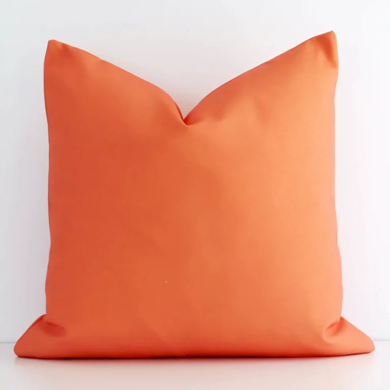 An attractive solid coloured outdoor cushion in front of a white brick wall. It has a square shape and is coral in colour.