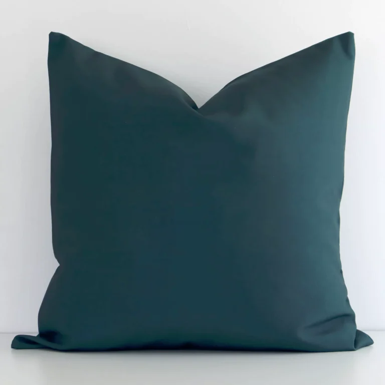 Dark green solid-colured cushion cover sits against a white wall. It is constructed from a superior looking linen material and has a square shape.