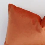 Extreme close up of a rectangle burnt orange cushion. The solid-coloured style and velvet fabric are shown with a much higher degree of detail.