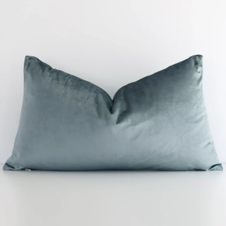 White wall with a solid-coloured sage green cushion laying against it. It has a distinctive velvet fabric and has a rectangle shape.