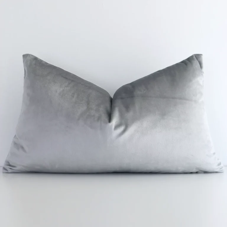 Velvet rectangle cushion with a solid-coloured design in an upright position against a white brick wall. It is silver in colour.
