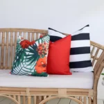 A rattan seat next to a green plant has 3 outdoor cushions from the Byron collection on it.