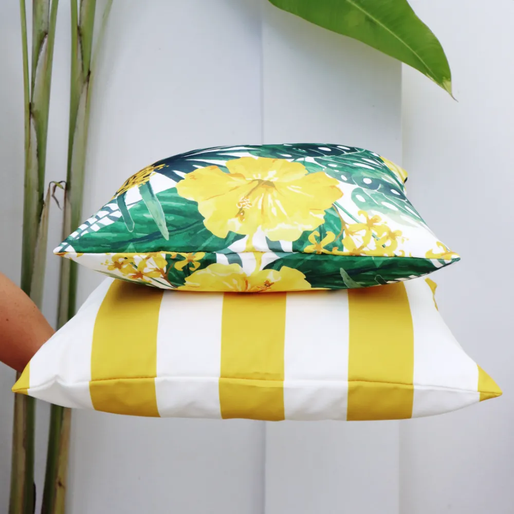 An arm is holding out an outdoor cushion set in front of a wall and plant.