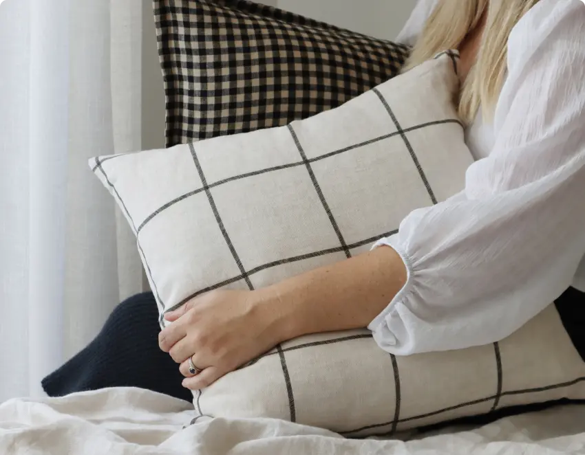 A woman is grasping two cushions whilst sitting on a bed.