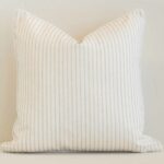 White wall with a stripe sand cushion laying against it. It has a distinctive outdoor fabric and has a square shape.