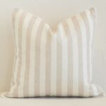 Square stripe cushion cover in sand colour sitting upright in front of a brick wall. It has been made from a quality outdoor material.