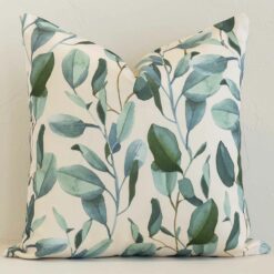 Floral cushion in cream and green colour positioned in front of a brick wall. It has square dimensions and is made from an outdoor material.