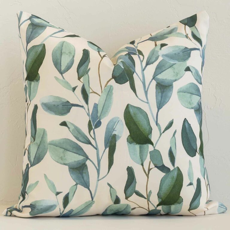 Floral cushion in cream and green colour positioned in front of a brick wall. It has square dimensions and is made from an outdoor material.
