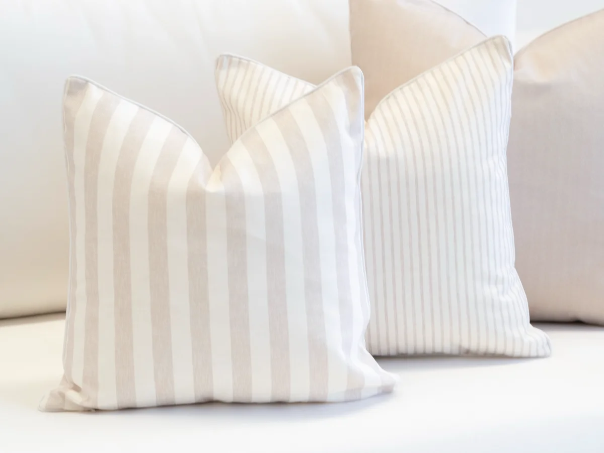 Three delicate sand beige coloured outdoor cushions in a pin stripe, regular stripe and a solid design are arranged on a seat.