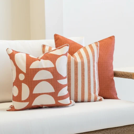 A set of 3 pacific rust outdoor cushions are arranged neatly in the corner of a white sofa.