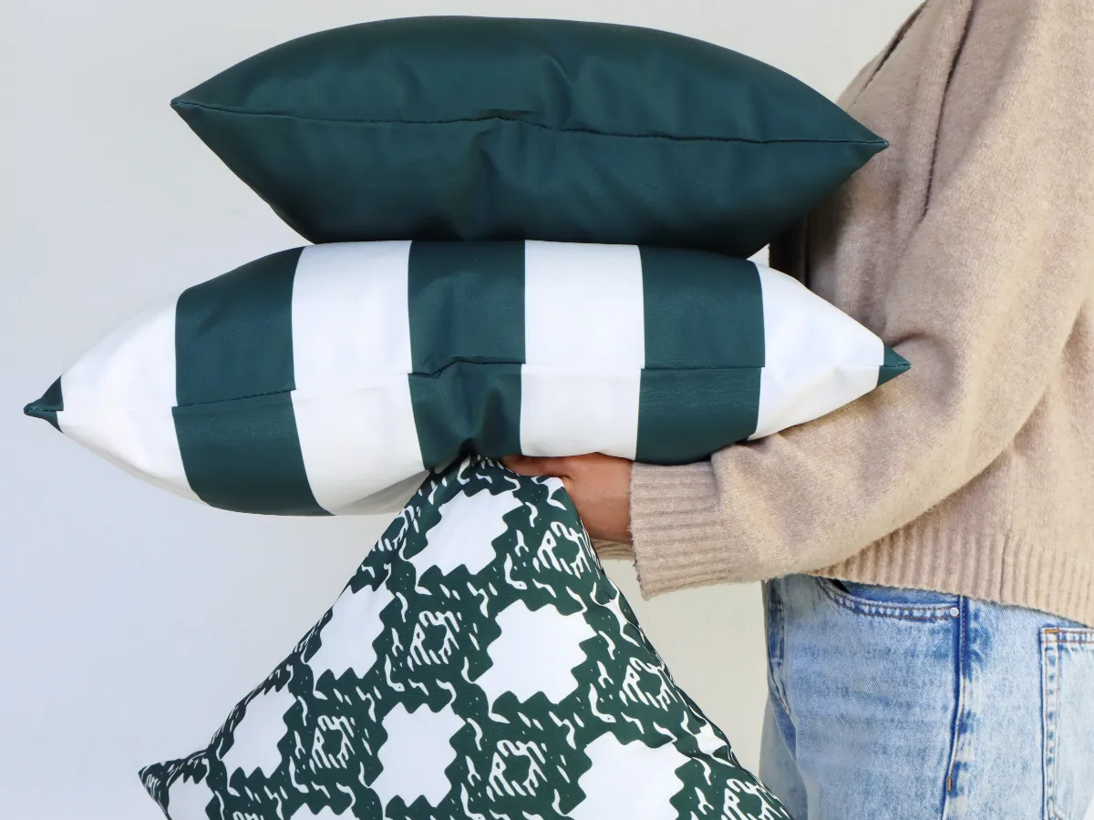 A woman is shown holding a stack of green outdoor cushions in front of a white external wall.