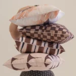 Person holding stacked Solterra cushions in warm brown and blush patterned designs.