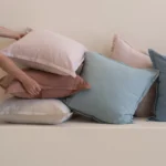 Hand placing pastel linen cushions stacked in soft blue, blush and rust tones.